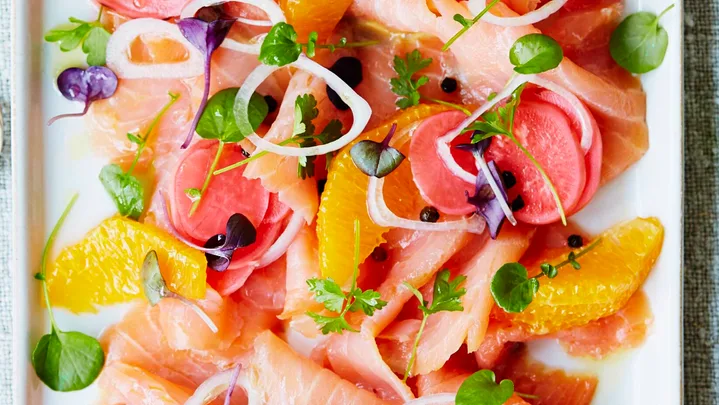 Plate of cured salmon with citrus, radish, microgreens, and onion rings, next to a stack of small forks on a linen cloth.