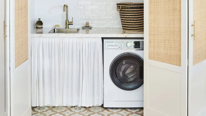 Laundry room with a washer, white curtain under sink, brass faucet, and a striped basket on patterned floor tiles.